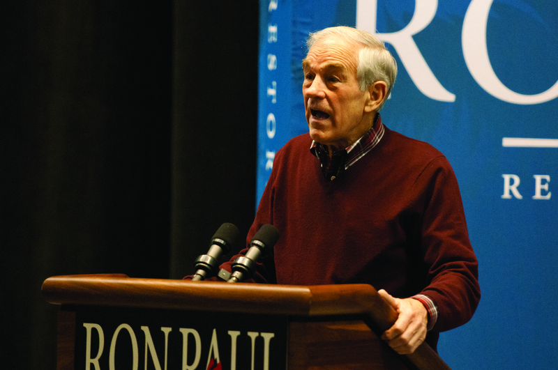 Color photograph of a white haired man in a brown sweater stands at a podium with the words "RON PAUL" on it.